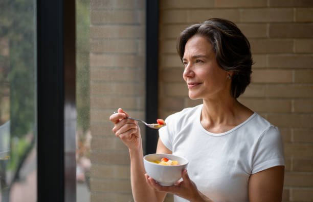 A woman stands by a large window holding a bowl of fruit, pausing mid-bite with a gentle smile, as she mindfully enjoys her food and gazes outside in soft natural light.